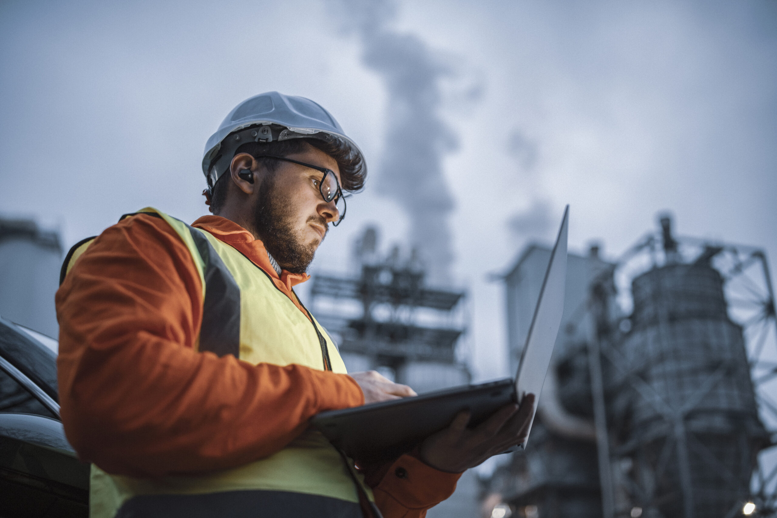 A shot of an young engineer wearing a helmet and using a laptop and hands free device during his night shirt in the oil rafinery. Engineering concept.